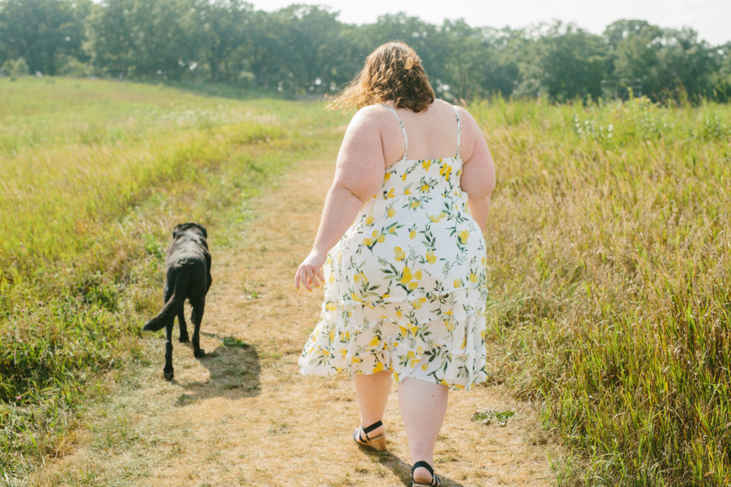 Kathy walks away from the camera along a grassy pathway in Glacial Park. She wears a white dress with lemons printed on it. Dexter the black dog walks alongside her with his tail mid wag. 