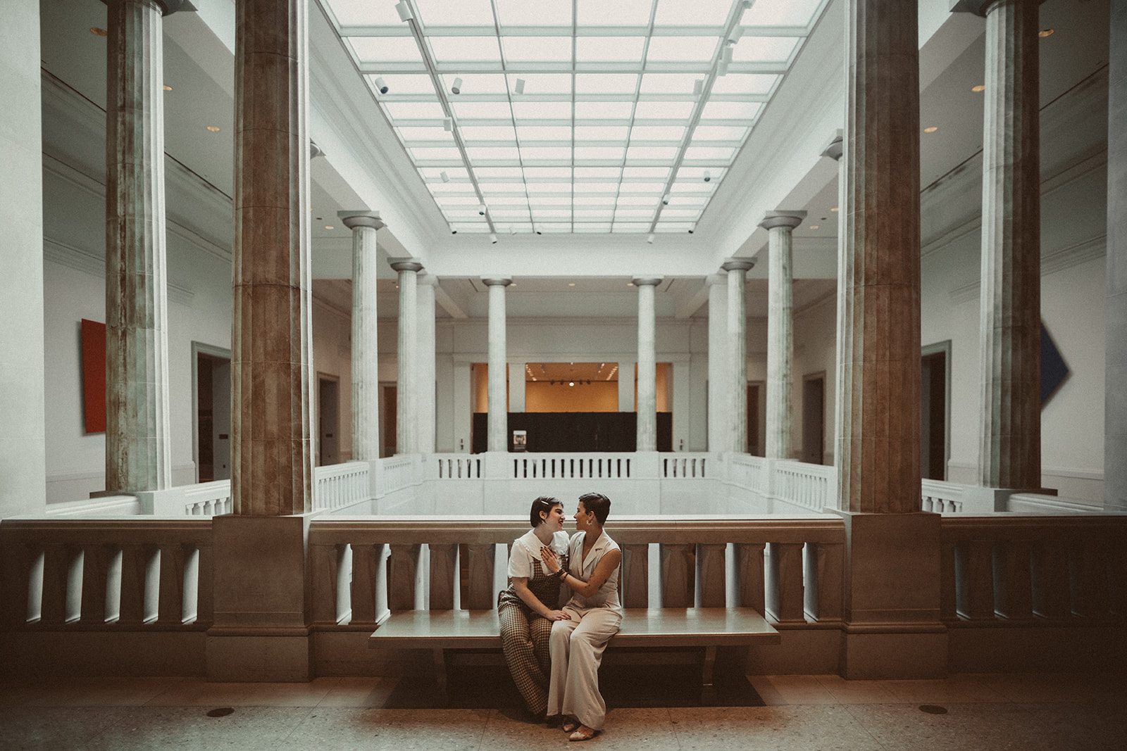 A grand room of columns and pillars with the couple sitting in the middle on the bench holding one another.