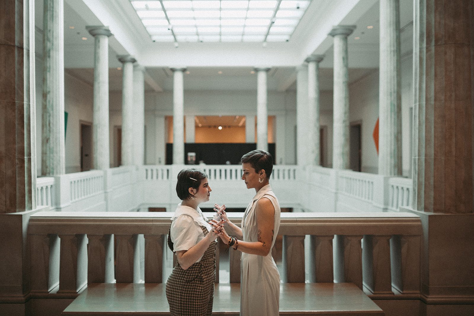 They face each other in front of a grand room of white marble pillars. They connect hands and stare at one another.