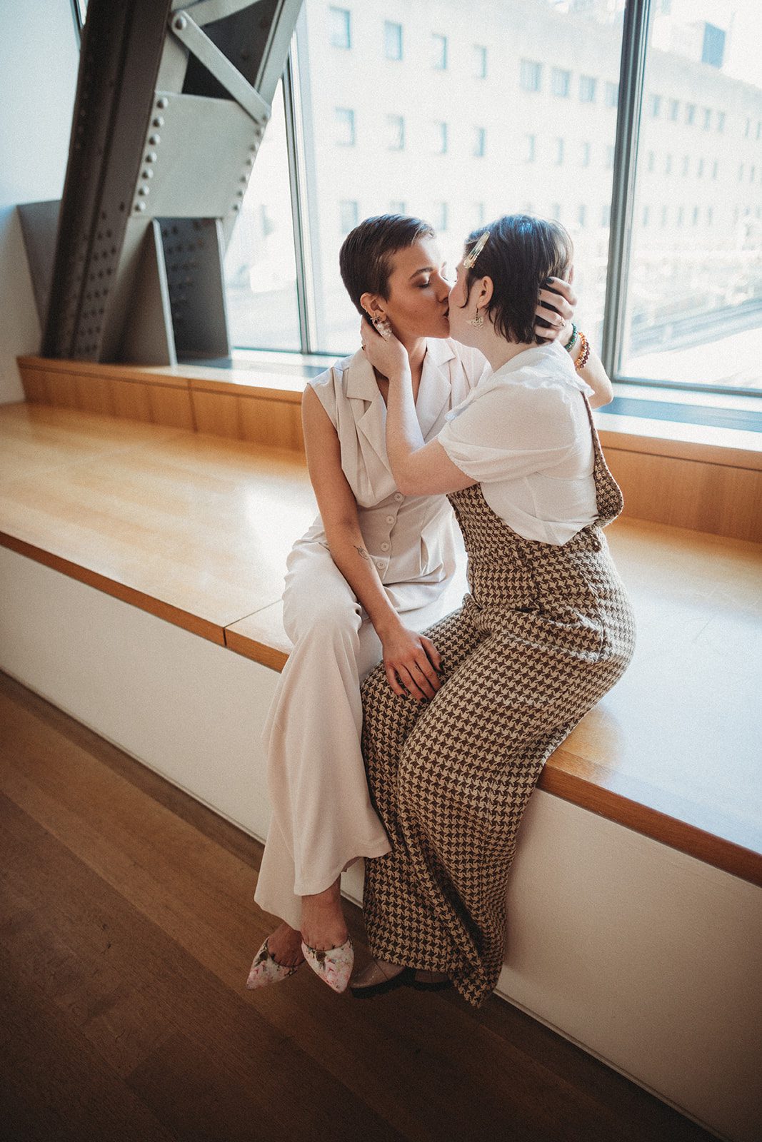 Sitting by the window, the Aves and Lex passionately kiss. They wear jumpsuits, Aves is cream colored and Lex wheres a cross stitched pattern with an overall top. Aves top is more formal, yet very elegant and modern. The both wear various jewelry and snappy shoes. 