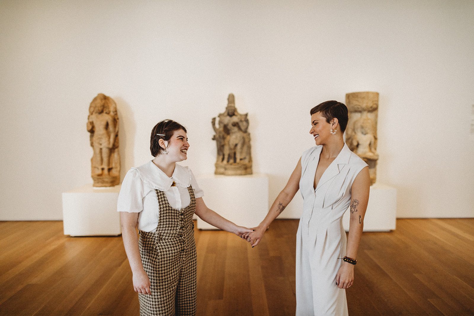 In front of an exhibit, the two outstretch their arms to hold hangs and smile at each other.